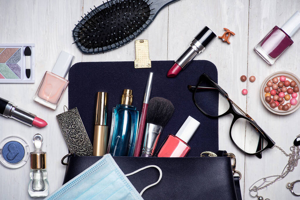 Mascarilla quirúrgica con varios productos de maquillaje y cosméticos que caen de una bolsa femenina mesa plana - Foto, Imagen