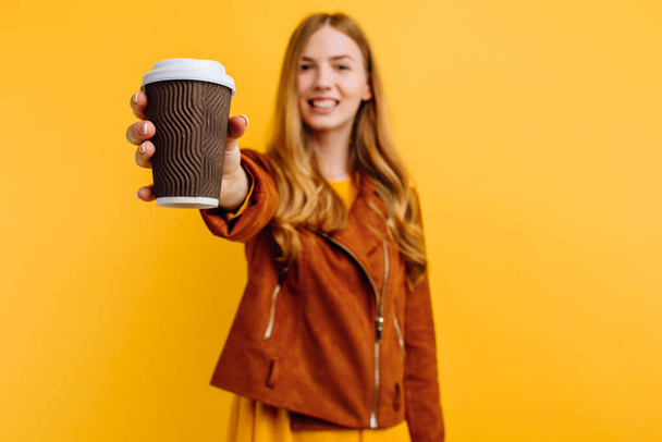 Stylish happy beautiful young woman in a yellow dress and autumn jacket, holding a paper Cup of coffee, on an isolated yellow background. The concept of autumn - Photo, image