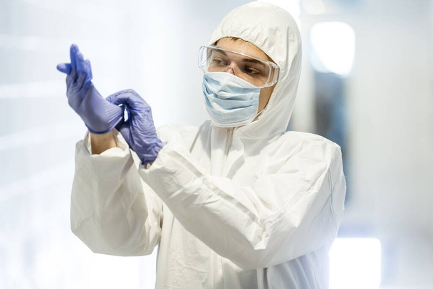 Corona virus concept. Male scientist doctor in mask, glasses and protective suit putting on latex gloves and getting ready for COVID-19 pandemic outbreak quarantine. Man in biochemistry laboratory. - Photo, image