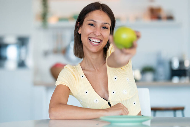 Portrait of beautiful young woman showing a green apple to the camera in the kitchen at home. - 写真・画像