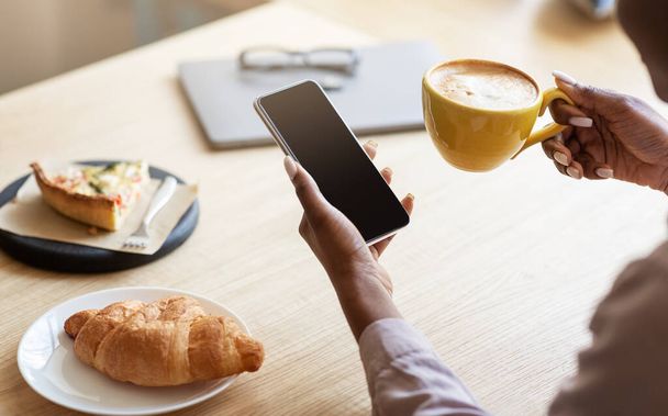 Femme noire méconnaissable utilisant un smartphone tout en prenant le petit déjeuner avec café au café, espace pour le design à l'écran - Photo, image