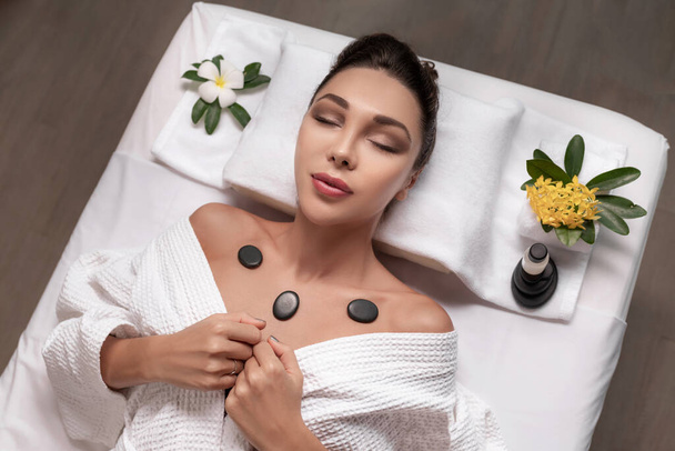 woman with closed eyes in white bathrobes relaxing on massage table with hot stones on her chest - Photo, image