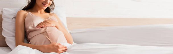 Waagerecht geschnittene Schwangere im seidenen Nachthemd berührt Bauch im Schlafzimmer  - Foto, Bild