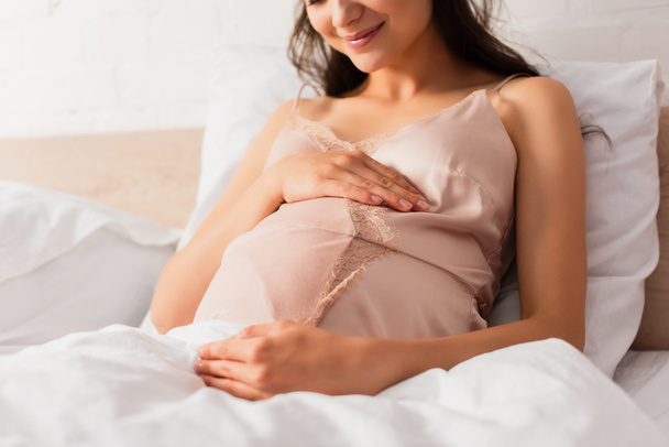 vista cortada da mulher grávida em seda nightie tocando barriga no quarto  - Foto, Imagem