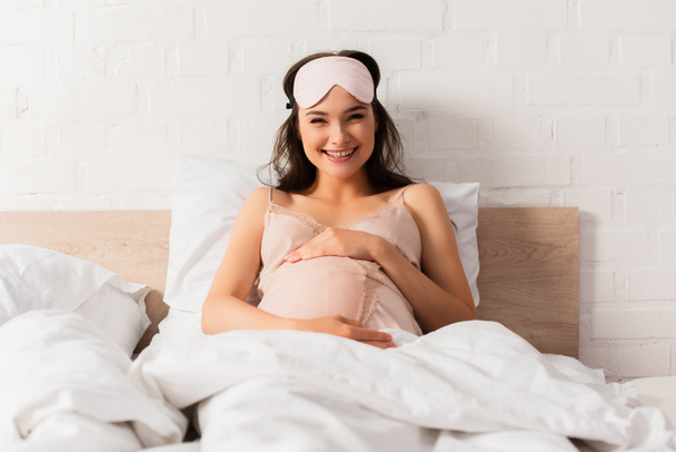 Schwangere junge Frau im seidenen Nachthemd berührt Bauch im Schlafzimmer  - Foto, Bild