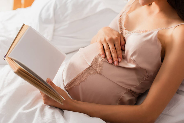 partial view of pregnant young woman in silk nightie holding book while touching belly in bedroom - Foto, afbeelding