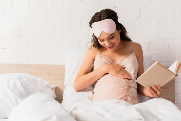 femme enceinte en nuisette de soie et masque pour les yeux tenant le livre et toucher le ventre dans la chambre - Photo, image