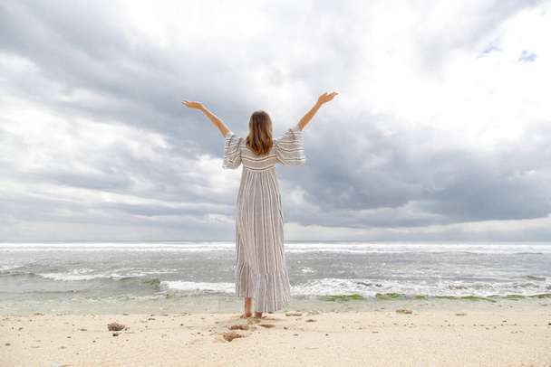 女性は腕を上げ、水平線を見てビーチに立っている。暗い夏の日の間に海岸沿いの長い白いドレスを着た女性のバックビュー。自由、幸福、肯定の概念. - 写真・画像