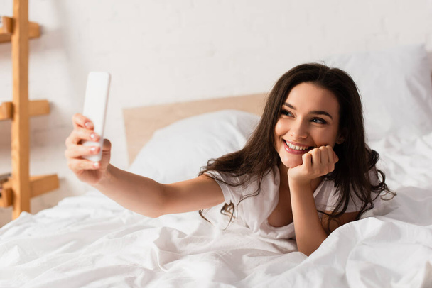 selective focus of young woman lying on bed and taking selfie  - Foto, afbeelding