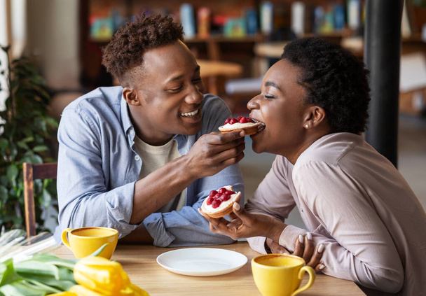 Millennial black man feeding tasty berry tartlet to his girlfriend at cozy cafe - Zdjęcie, obraz