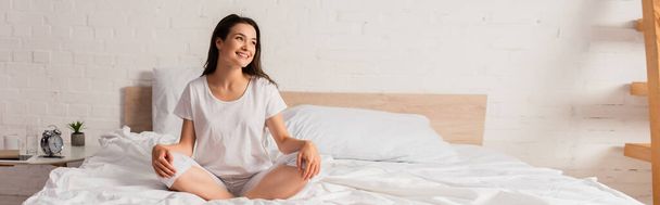 horyzontalny obraz brunetki siedzącej na łóżku i odwracającej wzrok  - Zdjęcie, obraz