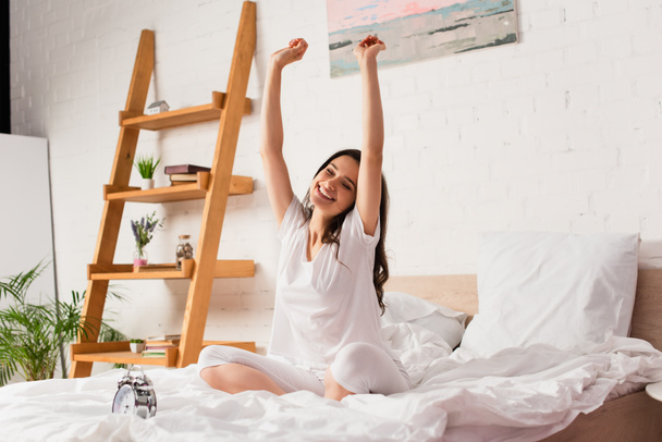 молодая женщина сидит на кровати и растягивается возле ретро будильника  - Фото, изображение
