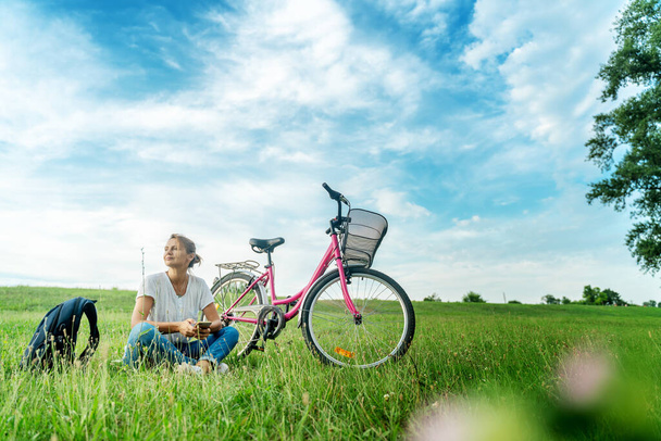 Beautiful smiling mature woman sitting next to bike on the grass on a green field. Summer Country Vacation and Adventure Concept - Fotoğraf, Görsel