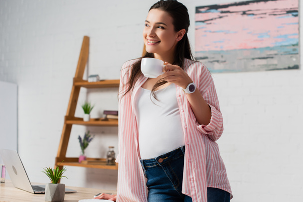 zwanger vrouw weg te kijken terwijl het houden van kopje thee terwijl staan in de buurt van laptop  - Foto, afbeelding