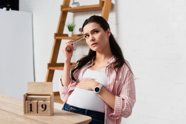 selective focus of thoughtful and pregnant woman touching belly near wooden cubes with date - Foto, imagen