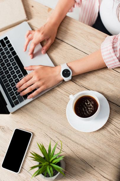 Ansicht von Freelancer mit Laptop in Kaffee-, Pflanzen- und Smartphone-Nähe mit leerem Bildschirm  - Foto, Bild