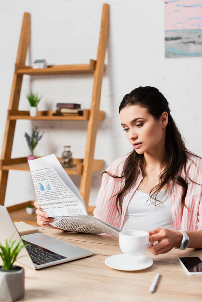 selectieve focus van zwangere vrouw het lezen van krant en holding cup in de buurt van gadgets  - Foto, afbeelding