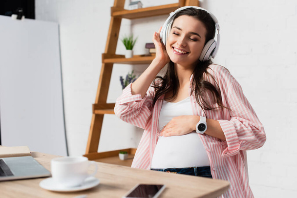 selektivní zaměření těhotné ženy se zavřenýma očima poslech hudby v bezdrátových sluchátkách v blízkosti gadgets a pohár  - Fotografie, Obrázek
