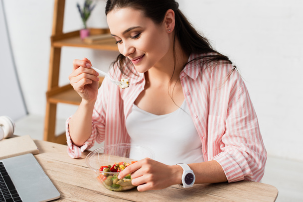 young brunette woman holding plastic fork with fresh salad near takeaway container  - Foto, imagen