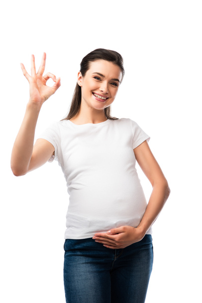 Schwangere im weißen T-Shirt berührt Bauch und zeigt Ok-Zeichen isoliert auf weißem Grund  - Foto, Bild