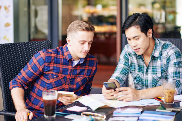 Jeunes multiethniques en chemises à carreaux se réunissant à la table du café et discutant d'une nouvelle application de voyage sur smartphone - Photo, image