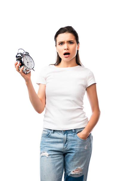 mujer conmocionada en camiseta blanca y jeans de pie con la mano en el bolsillo y la celebración de reloj despertador retro aislado en blanco - Foto, imagen
