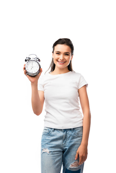 joven morena mujer en camiseta blanca y jeans con reloj despertador retro aislado en blanco - Foto, Imagen