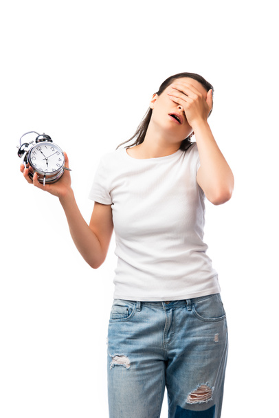 mujer cansada en camiseta blanca y pantalones vaqueros sosteniendo reloj despertador retro mientras cubre la cara aislada en blanco - Foto, imagen
