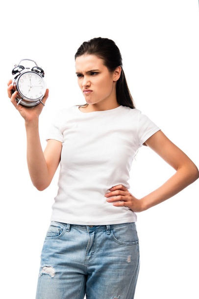 nespokojená žena v bílém tričku drží retro budík, zatímco stojí s rukou na boku izolované na bílém - Fotografie, Obrázek