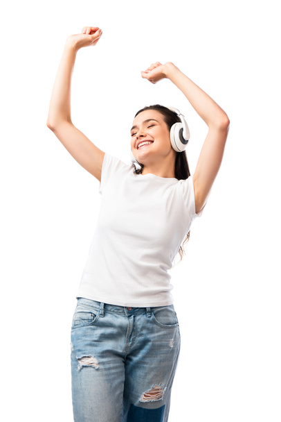 young woman in white t-shirt and wireless headphones dancing with hands above head isolated on white  - Fotografie, Obrázek