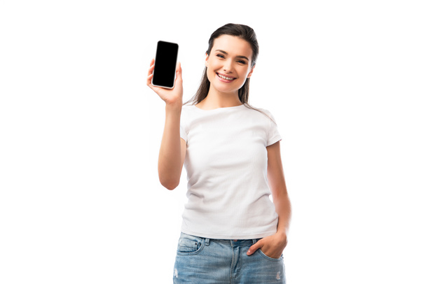 νεαρή γυναίκα σε λευκό t-shirt στέκεται με το χέρι στην τσέπη και κρατώντας smartphone με λευκή οθόνη απομονώνονται σε λευκό  - Φωτογραφία, εικόνα