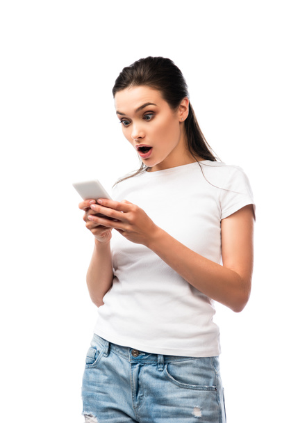 shocked brunette woman in white t-shirt using smartphone isolated on white - Fotoğraf, Görsel