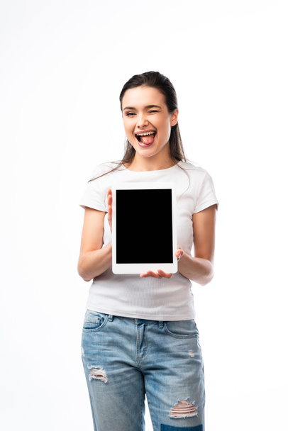 μελαχρινή γυναίκα σε λευκό t-shirt κρατώντας ψηφιακό δισκίο με λευκή οθόνη και να κολλήσει έξω γλώσσα απομονώνονται σε λευκό  - Φωτογραφία, εικόνα