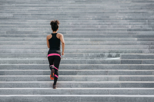 Spor giysili Afrikalı Amerikalı kaslı kız şehirde merdivenlerden koşarak çıkıyor. - Fotoğraf, Görsel