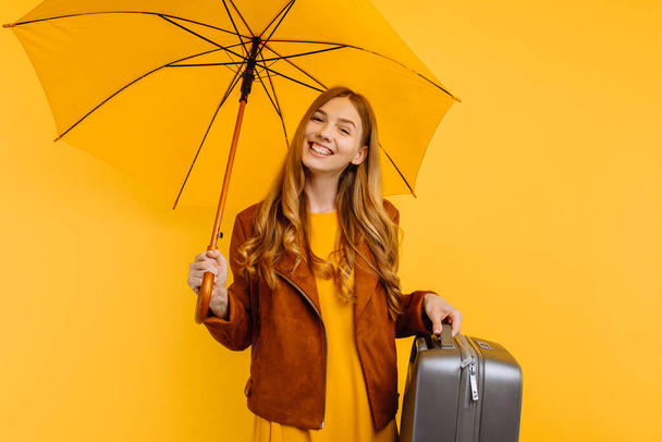 happy girl in a yellow dress and autumn jacket, standing with a suitcase and a yellow umbrella, on an isolated yellow background - Фото, изображение