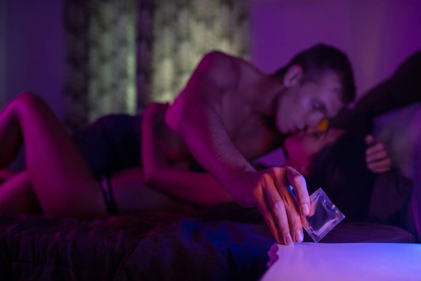 A guy kisses his girlfriend and reaches for a condom with his hand, in the foreground of which stands a condom on a bedside table near the lamp. Protected sexual intercourse. Love. Close-up. - Foto, Bild