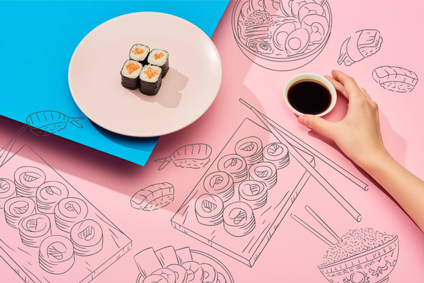 oříznutý pohled na ženu držící sójovou omáčku v blízkosti čerstvých maki s lososem a sushi ilustrací na modrém, růžovém pozadí - Fotografie, Obrázek