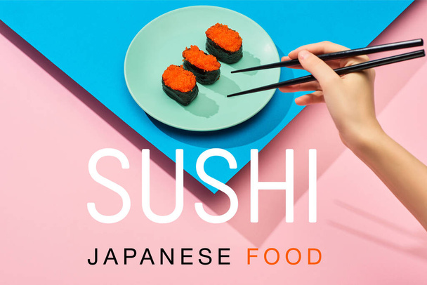 oříznutý pohled na ženu držící hůlky v blízkosti nigiri s červeným kaviárem a sushi japonské jídlo nápisy na modrém, růžovém pozadí - Fotografie, Obrázek