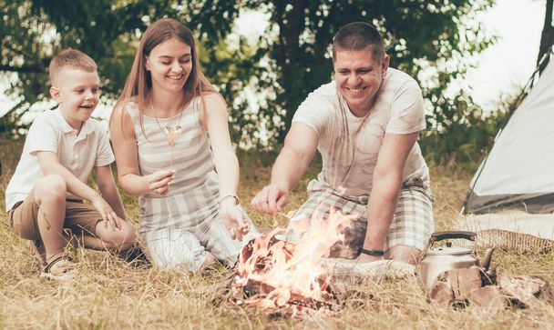  Uma família senta-se perto de uma tenda e uma fogueira e aquece marshmallows no fogo
 - Foto, Imagem