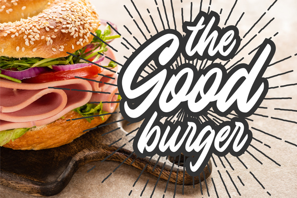delicious bagel with ham on wooden cutting board near the good burger lettering on textured surface - Fotó, kép