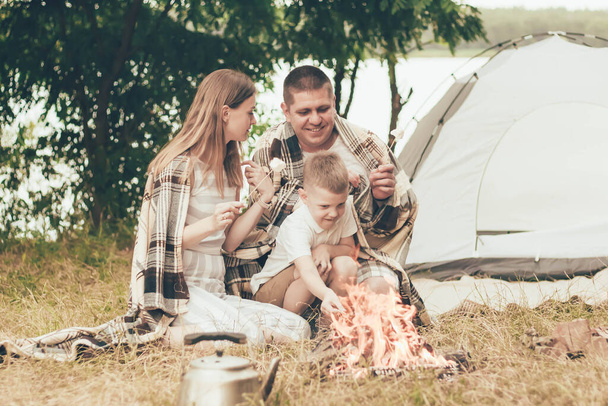 Mãe, pai e filho, cobertos com um cobertor, estão sentados perto da tenda e o fogo está aquecendo marshmallows no fogo
 - Foto, Imagem