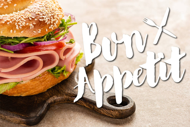 delicious bagel with ham on wooden cutting board near bon appetit lettering on textured surface - Photo, Image