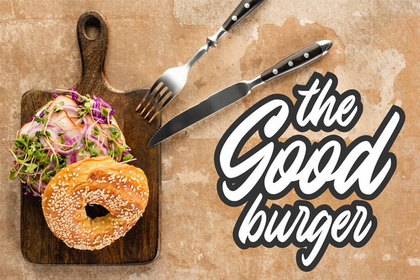 top view of fresh bagel with meat on cutting board with cutlery near the good burger lettering on textured surface  - Fotografie, Obrázek