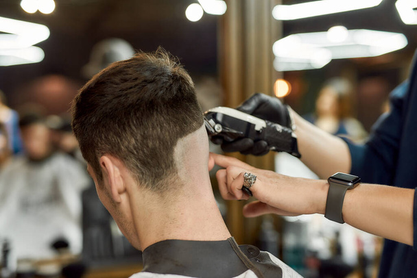 Visiting barbershop. Close up shot of professional barber working with hair clipper, making trendy haircut for client - Foto, afbeelding