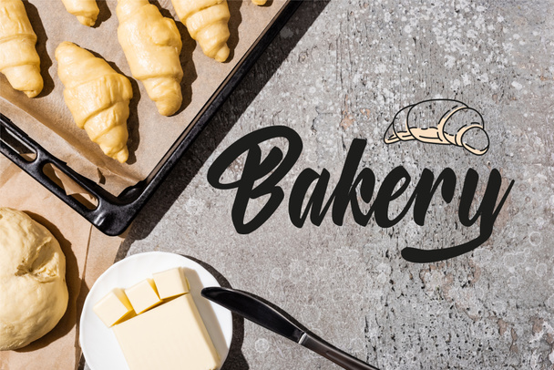 vista superior de croissants sin cocer en bandeja para hornear, masa y mantequilla cerca de letras de panadería en la superficie gris de hormigón - Foto, Imagen
