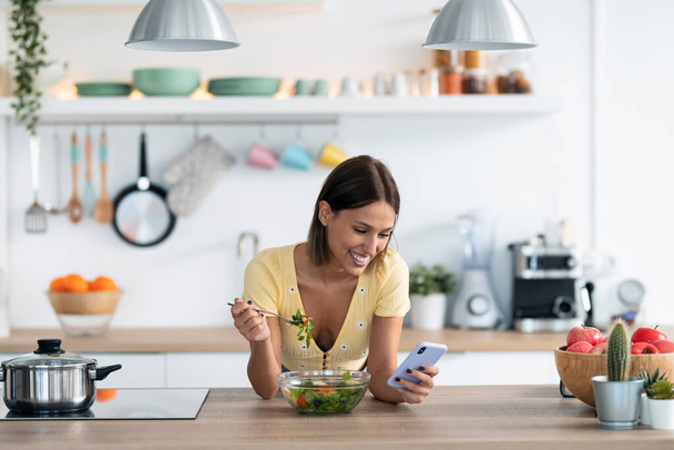Shot of beautiful young woman using her mobile phone while eating a salad in the kitchen at home.  - Фото, зображення