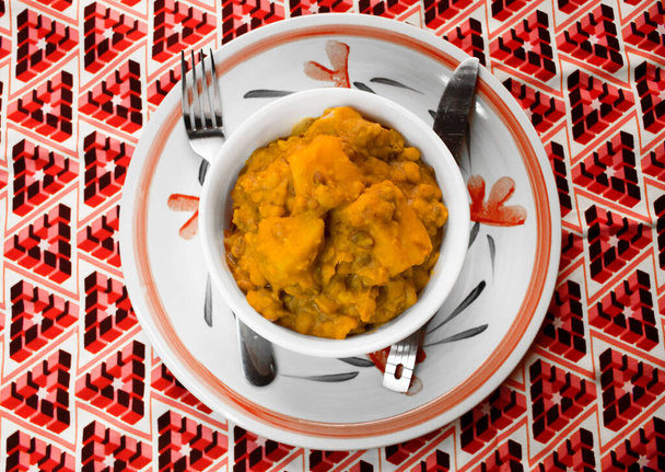 A white bowl of Yam and Beans Pottage or Nigerian Porridge served on a Plate with a colorful red pattern African tablecloth from top view - Photo, Image