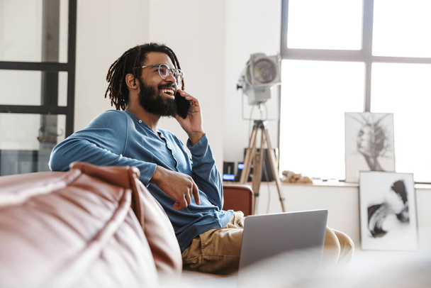 Schöner lächelnder junger afrikanischer Mann, der zu Hause auf einer Ledercouch sitzt, mit dem Handy telefoniert und Laptop benutzt - Foto, Bild