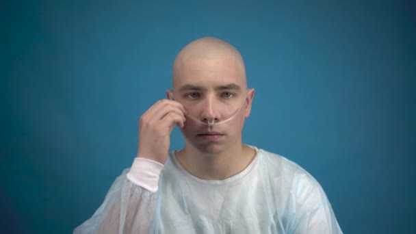 A bald young man with oncology sadly looks at the camera on a blue background. The patient straightens the tube of the breathing apparatus. Hair loss due to chemotherapy. - Footage, Video