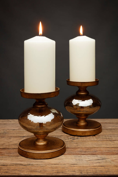 2 бронзовых подсвечника и светящиеся свечи, снятые на деревянном столе, на темно-сером фоне - Фото, изображение